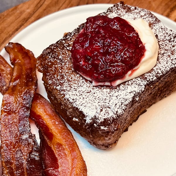 Thick French toast with powdered sugar, berry compote, whipped cream, and crispy bacon on a white plate.