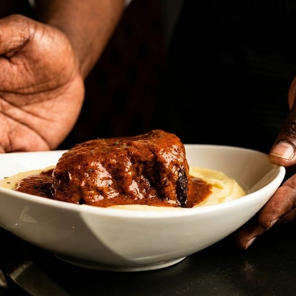 Hands presenting a white bowl of braised short rib with mashed potatoes and red wine.