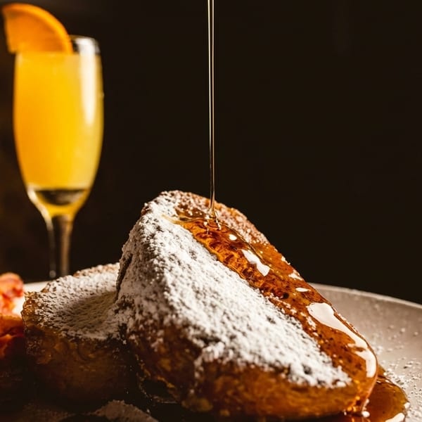 Maple syrup pouring over powdered sugar French toast with bacon and a mimosa.