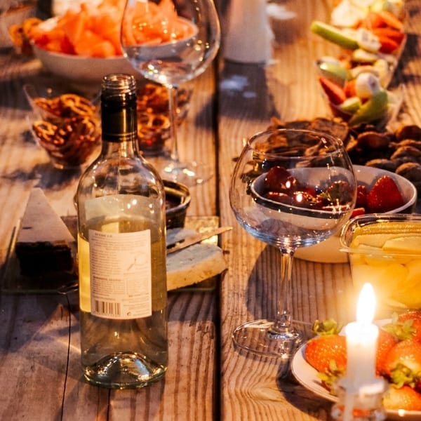A rustic wooden table set with wine, cheese, fruit, and snacks, illuminated by candlelight.