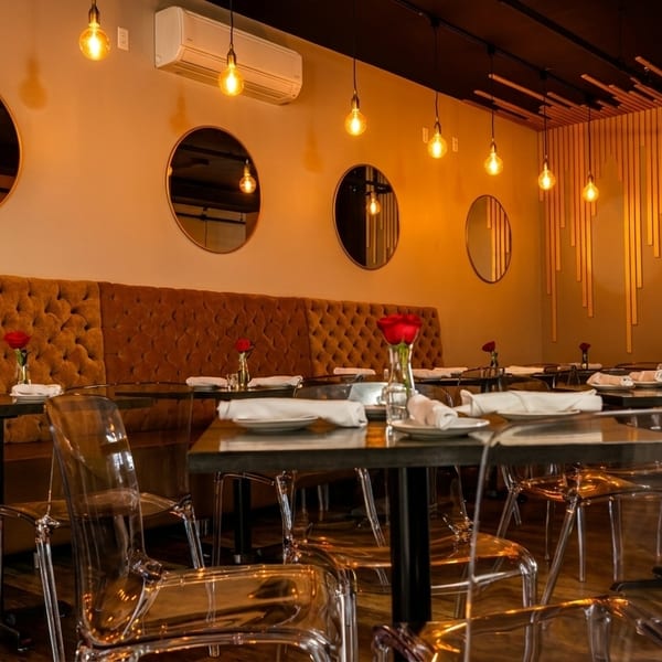 Modern restaurant interior with tufted orange banquettes, round mirrors, clear chairs, and pendant lights.