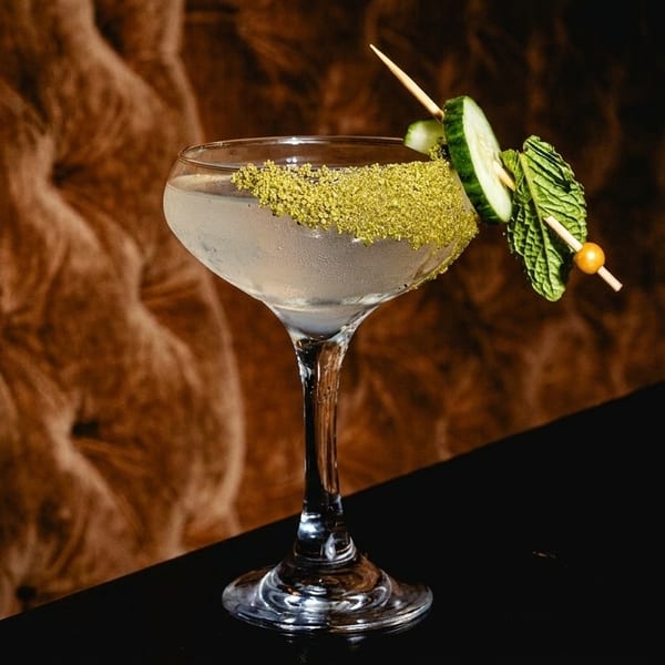 A cocktail in a coupe glass, garnished with green sugar, cucumber, and mint, on a dark surface.