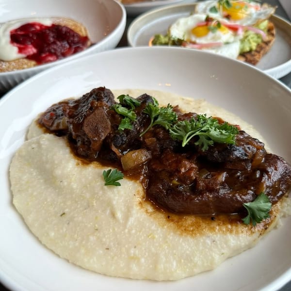 Braised short ribs with creamy grits, garnished with parsley, alongside other breakfast dishes.